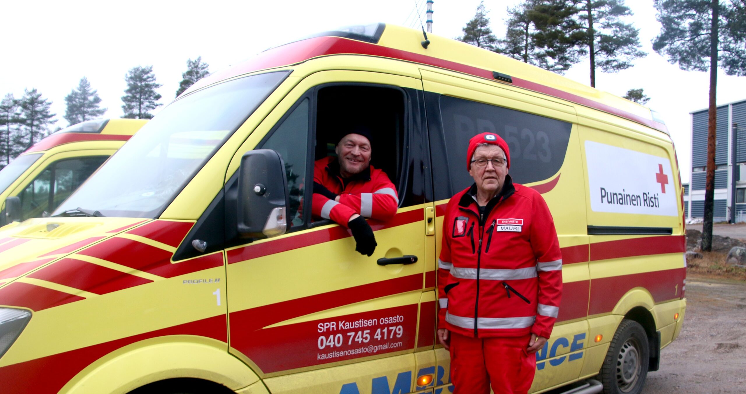 Kuvassa on SPR Kaustisen osaston ambulanssi. Kuskin penkillä istuu ensiapupäivystäjä Matti Mustajärvi ja auton vieressä seisoo ensiapupäivystäjä Mauri Koivusalo.