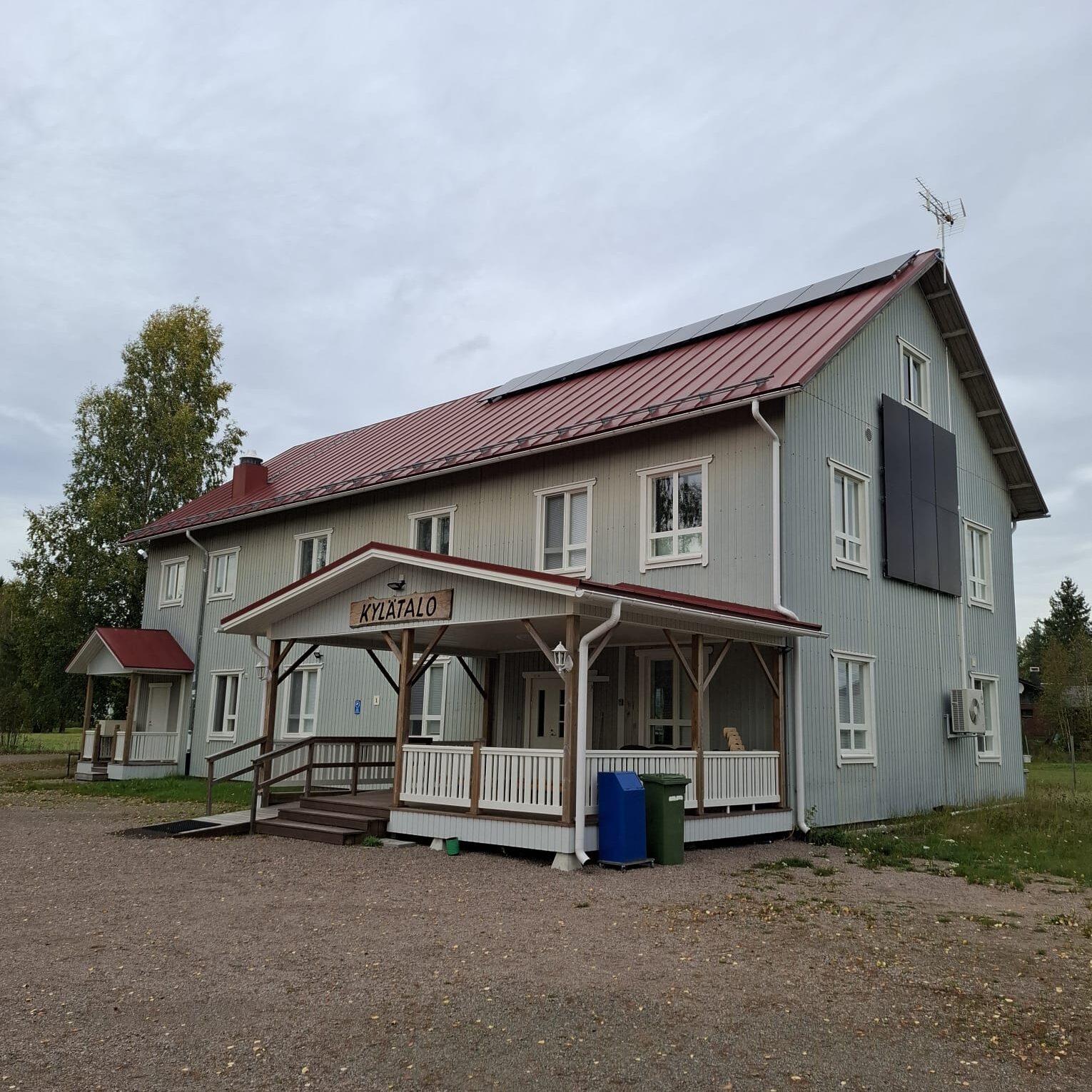 Kuvassa näkyy vaalea siniharmaa lautavuorinen, punakattoinen, 3-kerroksinen Halsuan Ylikylän kylätalo.